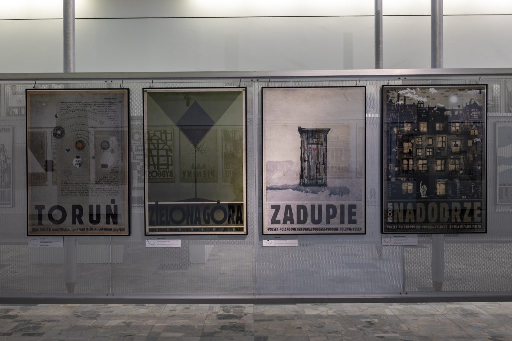 Widok sali wystawienniczej. Cztery plakaty przywieszone do metalowej ścianki wystawienniczej. Plakaty przedstawiają: Toruń, Zieloną Górę, Zadupie, Wrocław Nadodrze.